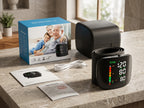 A digital wrist blood pressure monitor set displayed on a stone countertop, including the black cuff device, its blue-and-white retail box, a white cable, and printed paperwork. The background shows a softly blurred kitchen interior with a window.